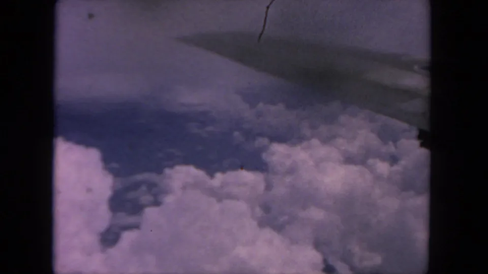 1962 home-movie frame from a passenger window, showing a wing edge over layered clouds and blue sky.