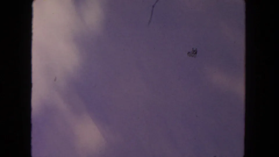 1962 8mm home-movie sky frame with a purple-blue cloud mass, pale cloud edge, dust marks, and film-gate borders.