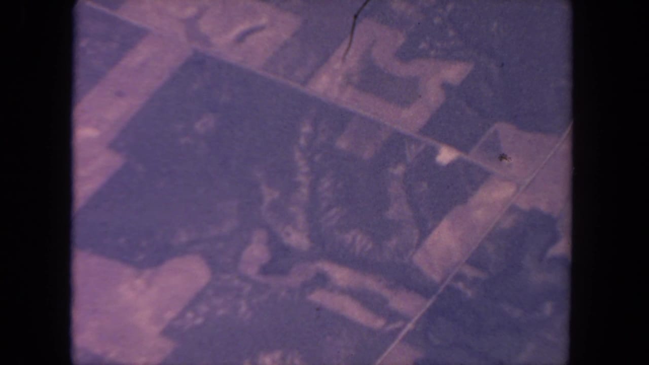 1962 San Diego aerial home-movie frame with geometric land parcels and dark irregular shapes under faded magenta and navy tones.