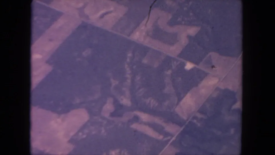 1962 San Diego aerial home-movie frame with geometric land parcels and dark irregular shapes under faded magenta and navy tones.