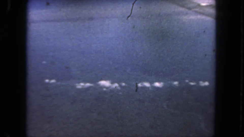 Grainy 1962 home-movie frame with a blue-gray moving surface, gate edges, dust, and scattered white reflections or marks.