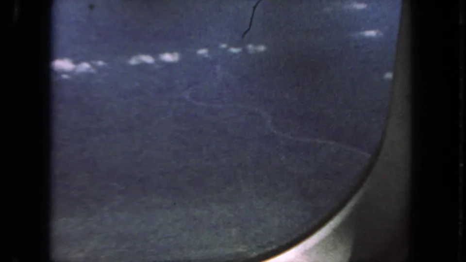 Grainy 1962 8mm frame of scattered clouds and blue sky viewed from inside an airplane window.