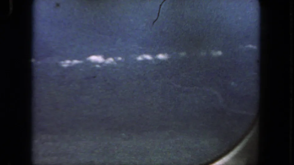 1962 home-movie still showing white cloud clusters across blue sky through a passenger airplane window.