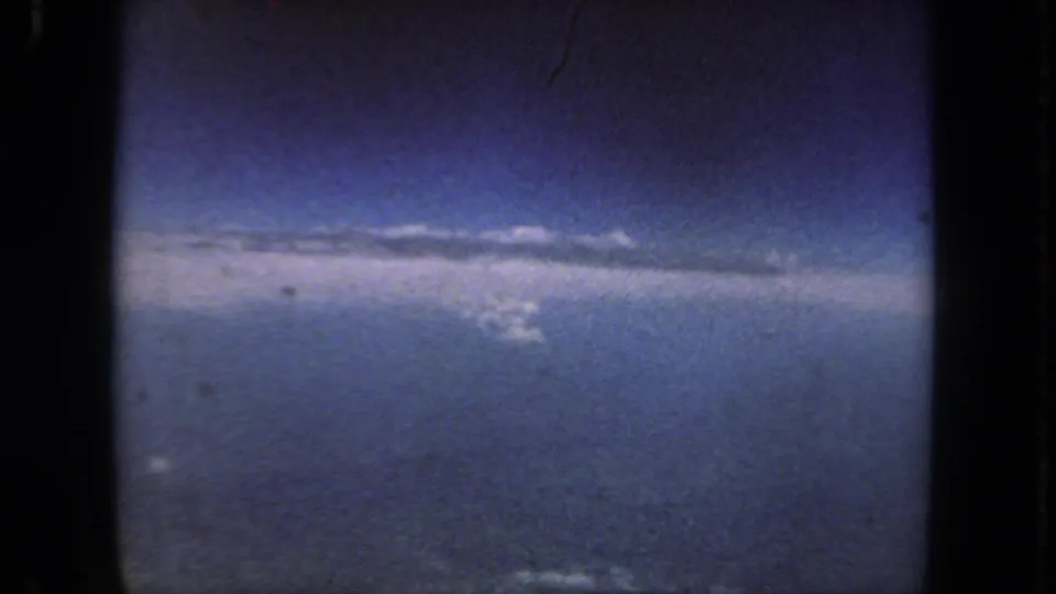 1962 home-movie airplane-window view with blue sky, a horizontal cloud band, and ground far below.