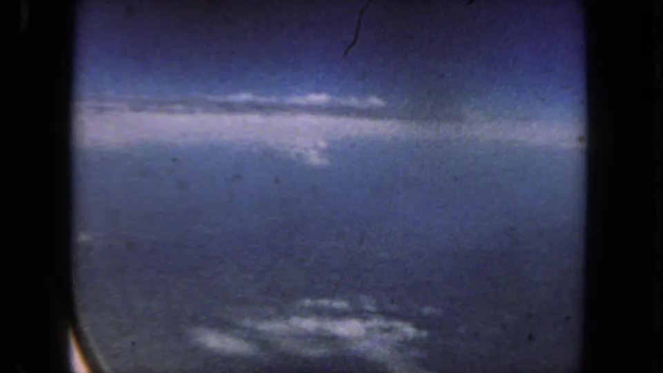 Vintage 8mm airplane-window shot showing scattered clouds, blue sky, and distant ground during flight.