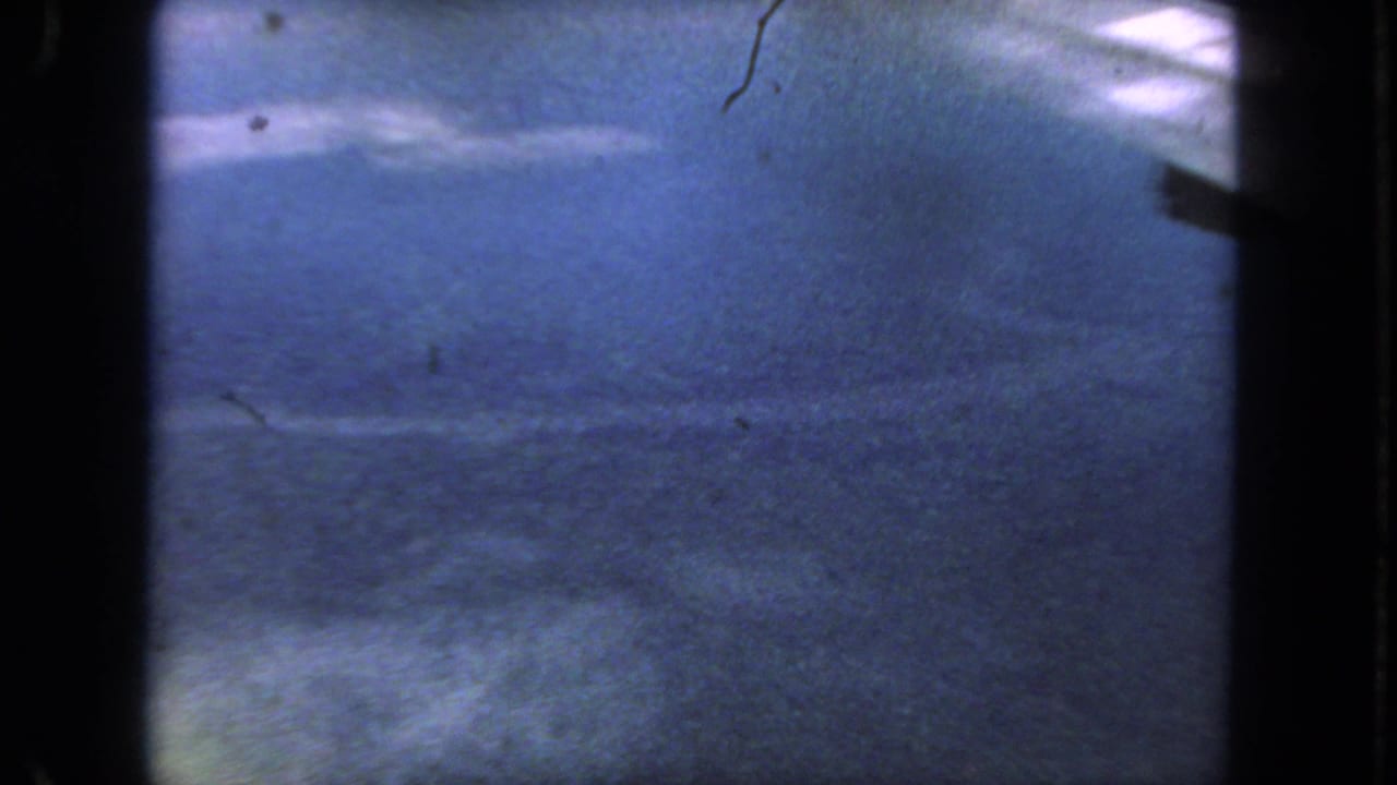 1962 San Diego 8mm frame showing blue-tinted road surface from a moving vehicle, bordered by dark film edges and dust specks.