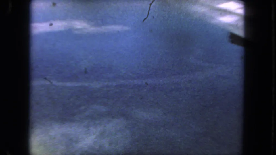 1962 San Diego 8mm frame showing blue-tinted road surface from a moving vehicle, bordered by dark film edges and dust specks.