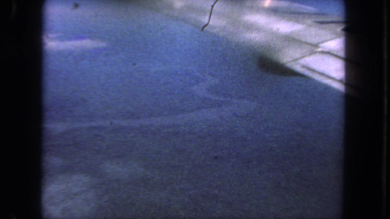 1962 San Diego home-movie frame with blurred pavement, a pale road edge, dark frame borders, and small film marks.