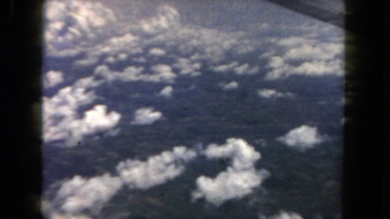 1962 home-movie frame showing clouds below and dark window borders from an airplane passenger view.
