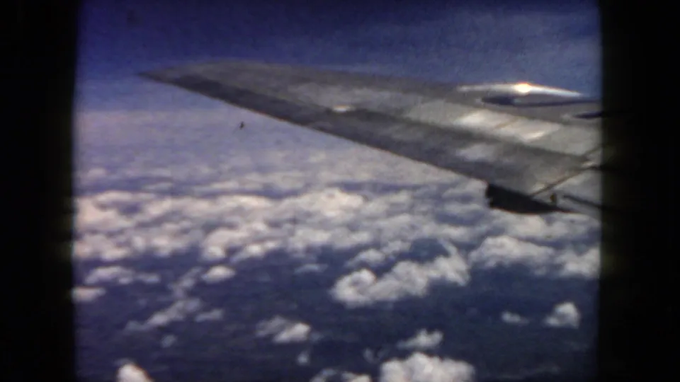 1962 8mm passenger view with an airplane wing stretching over scattered clouds and darker terrain or water.