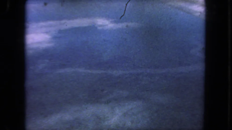 Faded 1962 8mm aerial frame with blue haze, faint cloud forms, and the edge of the airplane window.