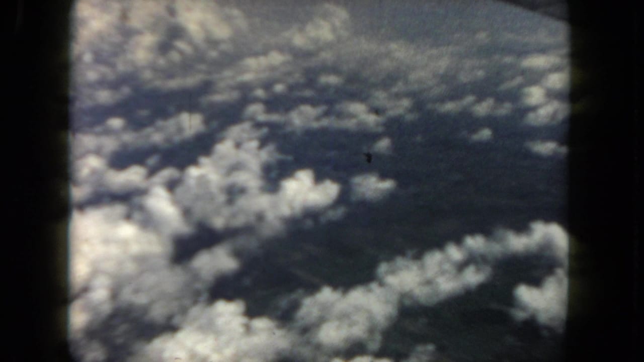 1962 8mm airplane-window view of scattered clouds over dark land or water, with soft focus and visible film borders.