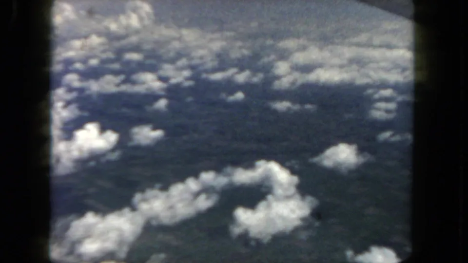 A 1962 home-movie frame looks down from an aircraft at scattered white clouds over dark land or water.