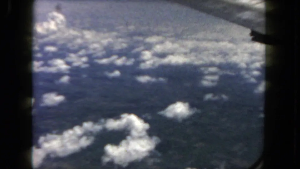 A partial airplane wing appears at the upper right over scattered clouds in this 1962 8mm aerial travel view.