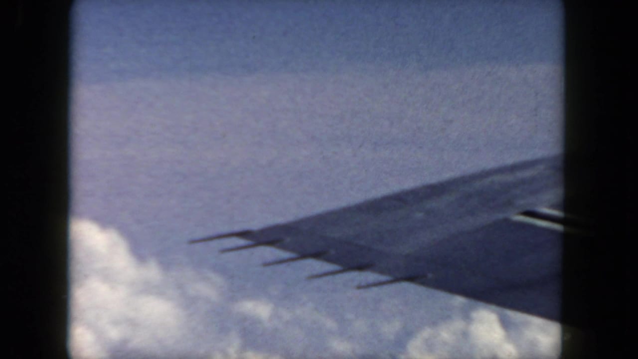 1962 aerial home-movie view with an airplane wingtip crossing blue sky above a bank of clouds.
