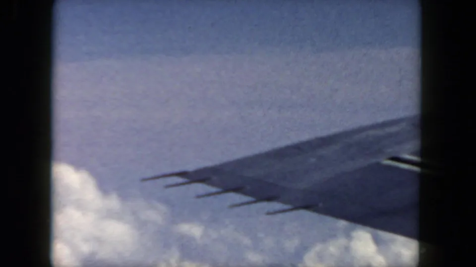 1962 aerial home-movie view with an airplane wingtip crossing blue sky above a bank of clouds.