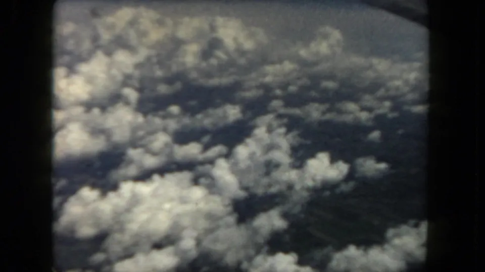 1962 8mm passenger-window view looking down over clouds and hazy terrain, with dark film or window borders visible.