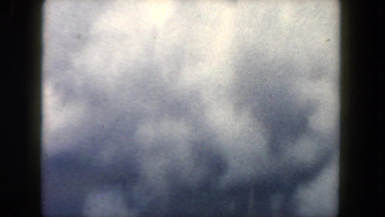 1962 in-flight home-movie view with cloud layers and a partial airplane wing visible beyond the dark window frame.