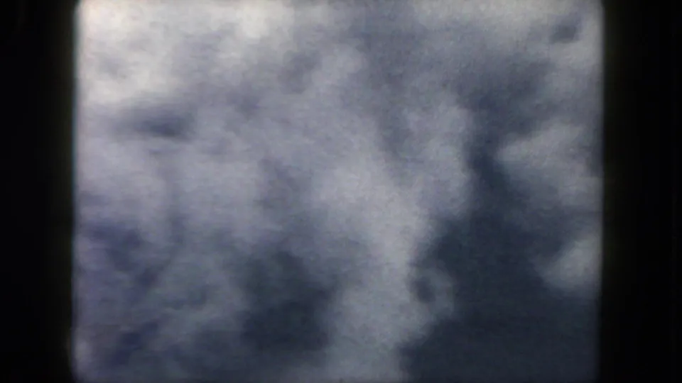 1962 home-movie aerial frame showing blurred clouds and shadowed land forms below, rendered with grain and blue-gray exposure.