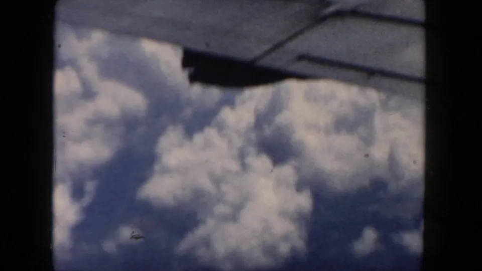 Passenger-window view from an airplane, with a dark wing edge crossing above sunlit clouds in 1962 home-movie footage.