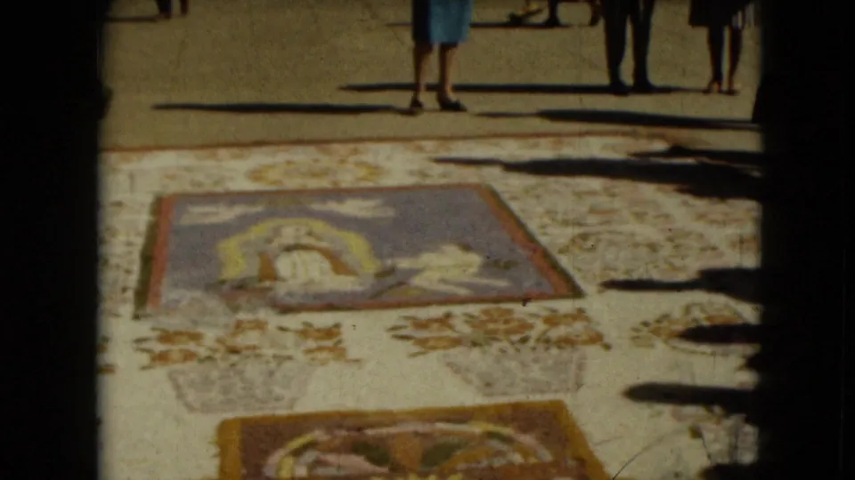 Mexico City 1962 home-movie frame with passersby partially visible above religious textile panels and long shadows on pavement.