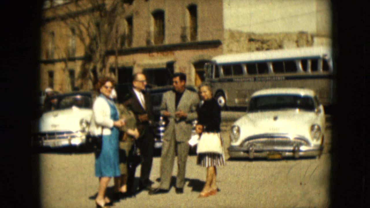In Mexico City archival 8mm footage from 1962, four adults converse near parked mid-century cars and a bus on a bright street.