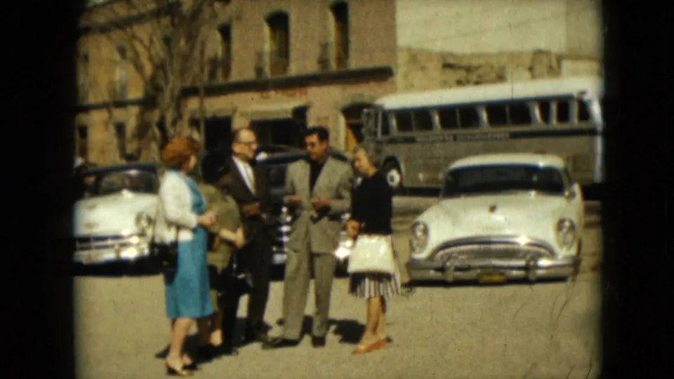 A 1962 Mexico City street scene shows adults in mid-century dress talking in front of parked cars and a bus.