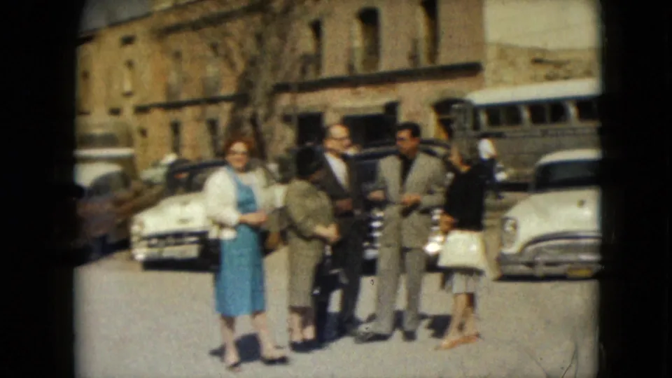 A woman in a blue dress faces the camera while others converse nearby on a 1962 Mexico City street with parked cars behind them.