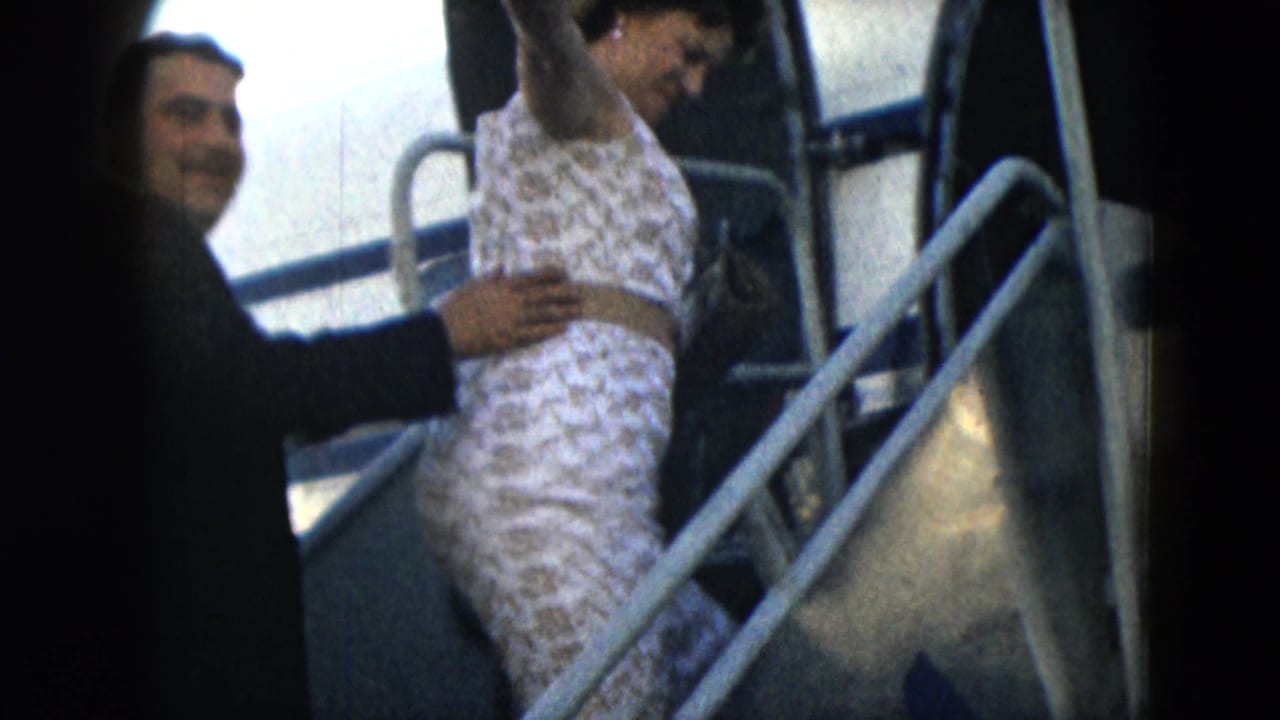 Man in dark suit helps a woman in a patterned white dress board aircraft stairs.