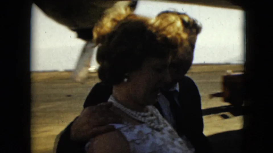 1962 Mexico City home-movie frame showing a formally dressed couple walking beside a propeller aircraft on the tarmac.