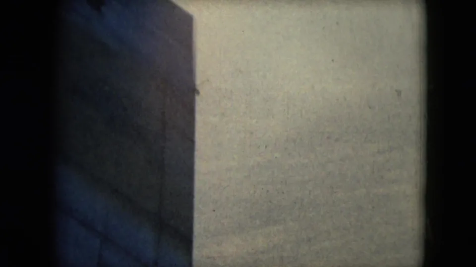 A soft 8mm frame shows mostly a pale building wall and dark frame edge before the couple enters view.