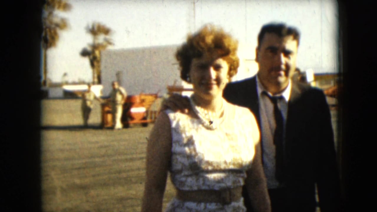 In Mexico City, 1962, a woman in a white lace dress and a man in a dark suit walk toward the camera outdoors.