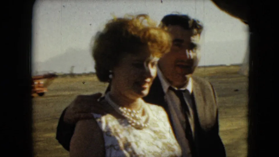 The formally dressed couple moves past the camera in Mexico City, with a broad paved area and distant buildings behind them.