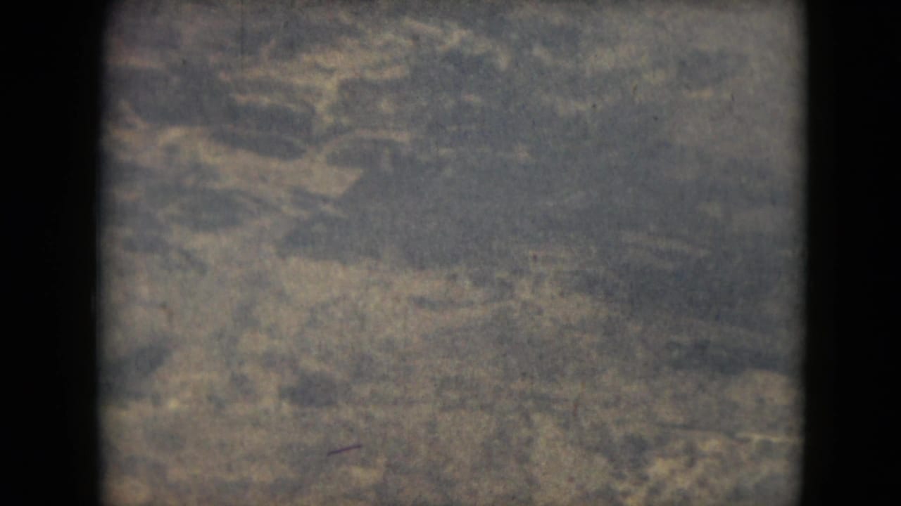 A grainy 1962 8mm frame shows distant ground texture from above, with no visible landmarks, people, or structures identifiable.