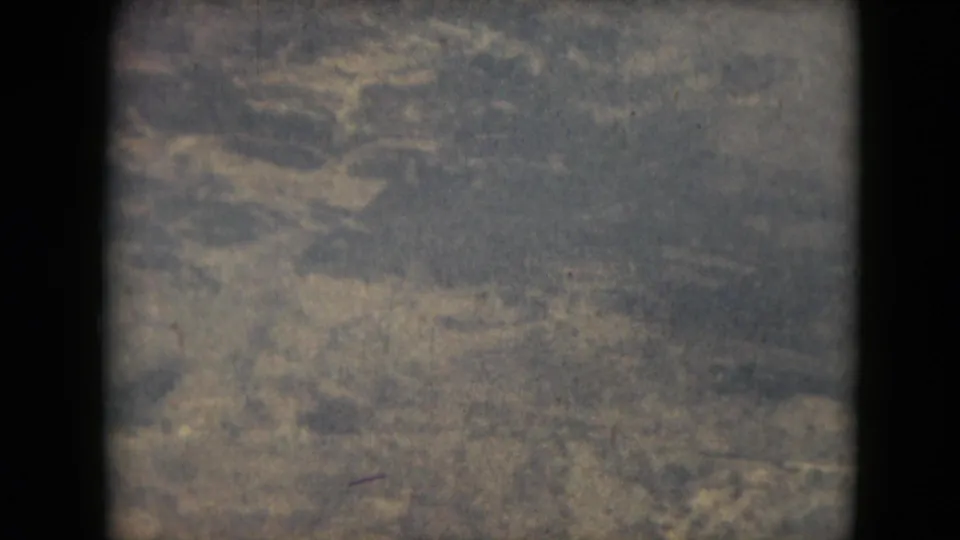 A grainy 1962 8mm frame shows distant ground texture from above, with no visible landmarks, people, or structures identifiable.