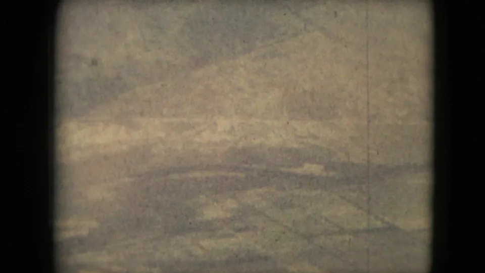 1962 8mm aerial frame with soft green-brown land patterns and faint cloud or haze, consistent with a traveler filming from an aircraft.