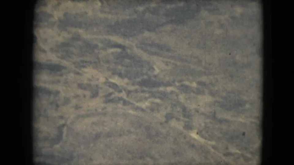 A warm, grainy 8mm aerial view shows ground textures and faint linear features from the airplane.