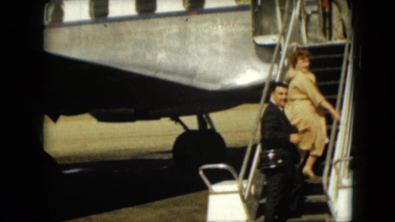 Woman climbs aircraft stairs while a man in a dark suit looks toward the camera.