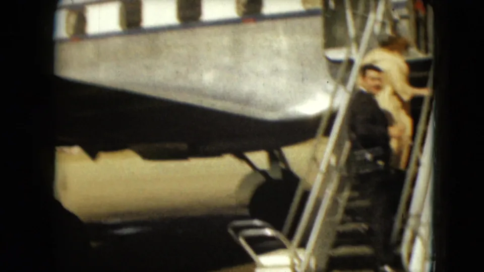 The handheld 8mm view shows the suited man on mobile aircraft stairs with the woman ahead near the plane doorway.