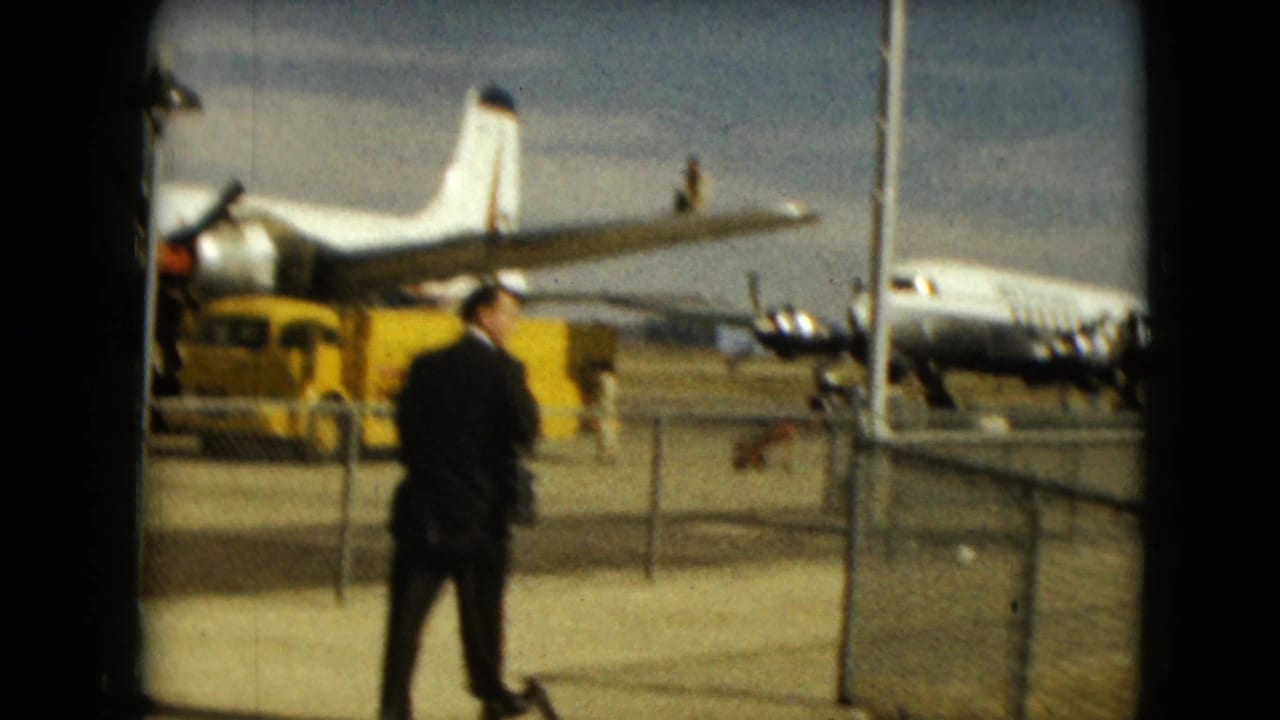 1962 Mexico City airport tarmac view with a suited man, distant propeller planes, fencing, and a yellow service vehicle.