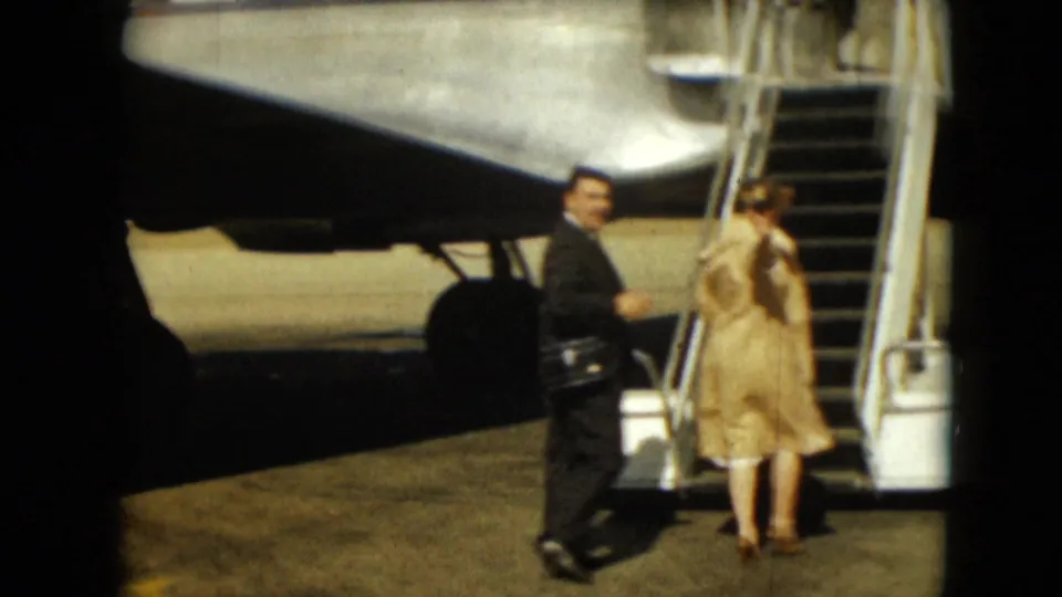 At the aircraft stairs, the woman faces the steps while the suited man looks back across the tarmac.