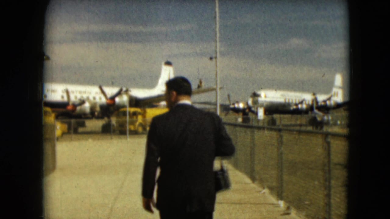 Suited man seen from behind walking along an airport path beside parked propeller planes.