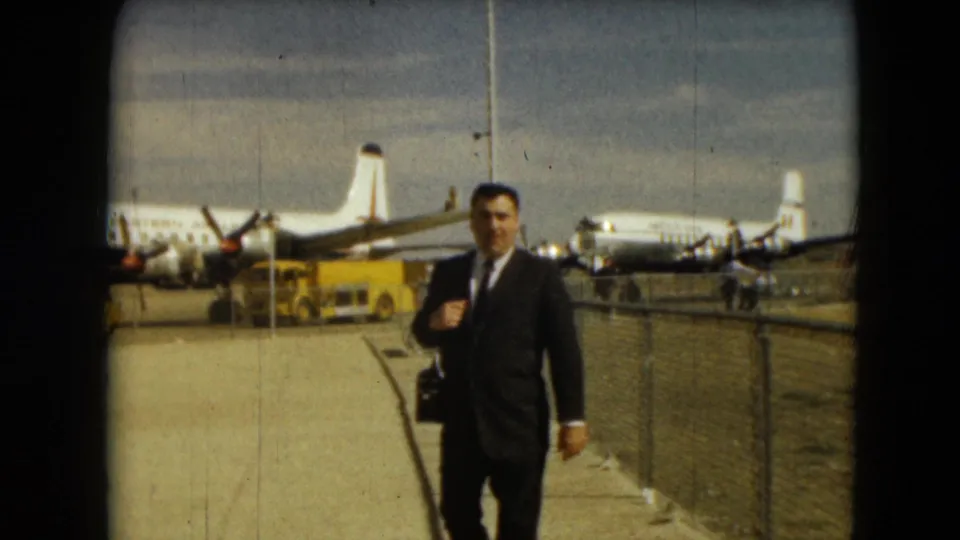 A suited traveler in Mexico City in 1962 approaches the camera with a black bag, with parked propeller planes and airport fencing behind him.