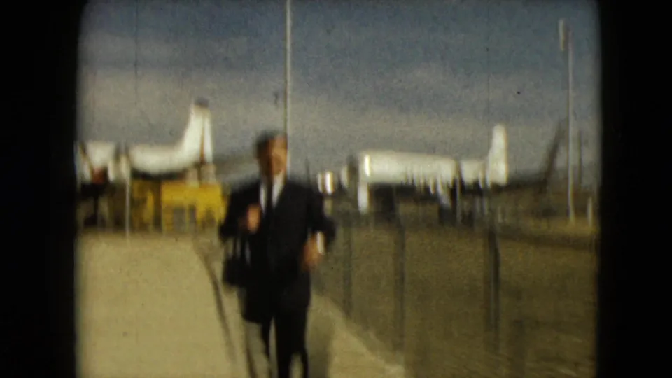 A soft-focused 1962 Mexico City airport frame shows the suited man moving close to the camera, with aircraft and fencing in the background.