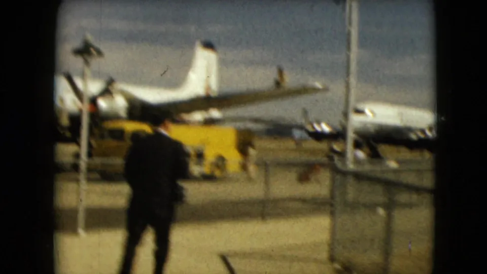At a Mexico City airport in 1962, the suited man walks away from the camera near a yellow service vehicle and a parked propeller aircraft.