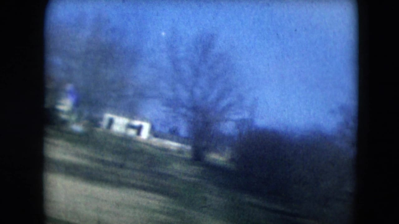Houses, trees, and open roadside land filmed through a car window.