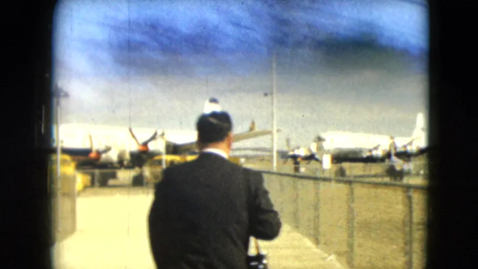1962 Mexico City airport scene showing a suited man from behind near a fence and parked propeller aircraft.