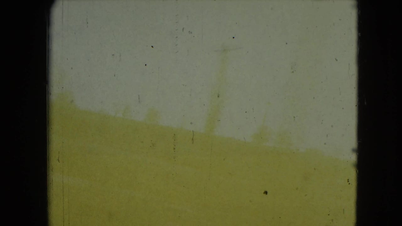 Yellowed 8mm frame from the moving car view, with indistinct outdoor shapes and dark window or gate edges.