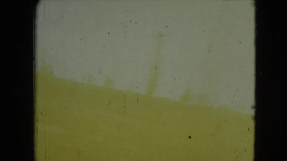 Yellowed 8mm frame from the moving car view, with indistinct outdoor shapes and dark window or gate edges.