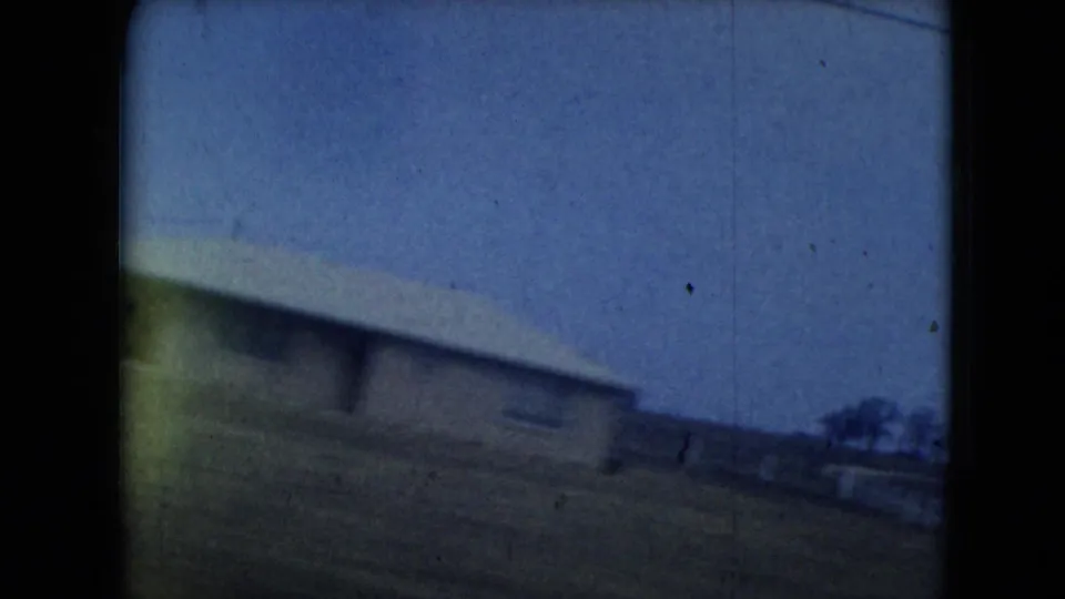 Blue-toned 1962 8mm roadside view with a low building, open ground, distant trees, and motion blur from a car window.